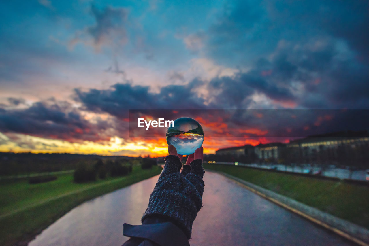 Cropped image of person holding crystal ball over river against cloudy sky