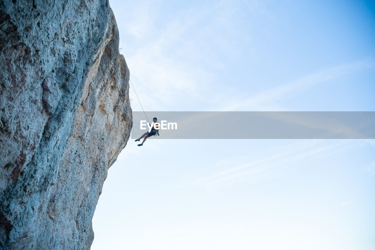 LOW ANGLE VIEW OF MAN CLIMBING ROCK ON CLIFF AGAINST SKY