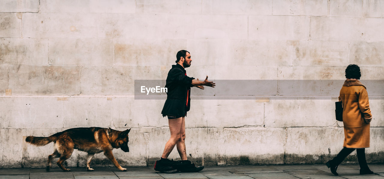 Side view of man with pants down walking by dog and woman against wall