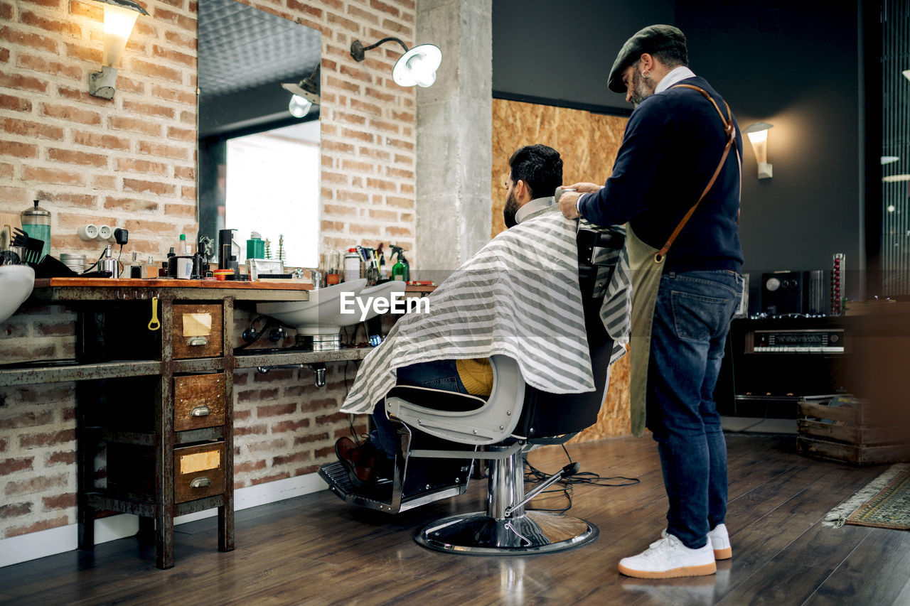 Back view male barber with comb brushing hair of unrecognizable client while making hairstyle in modern barbershop