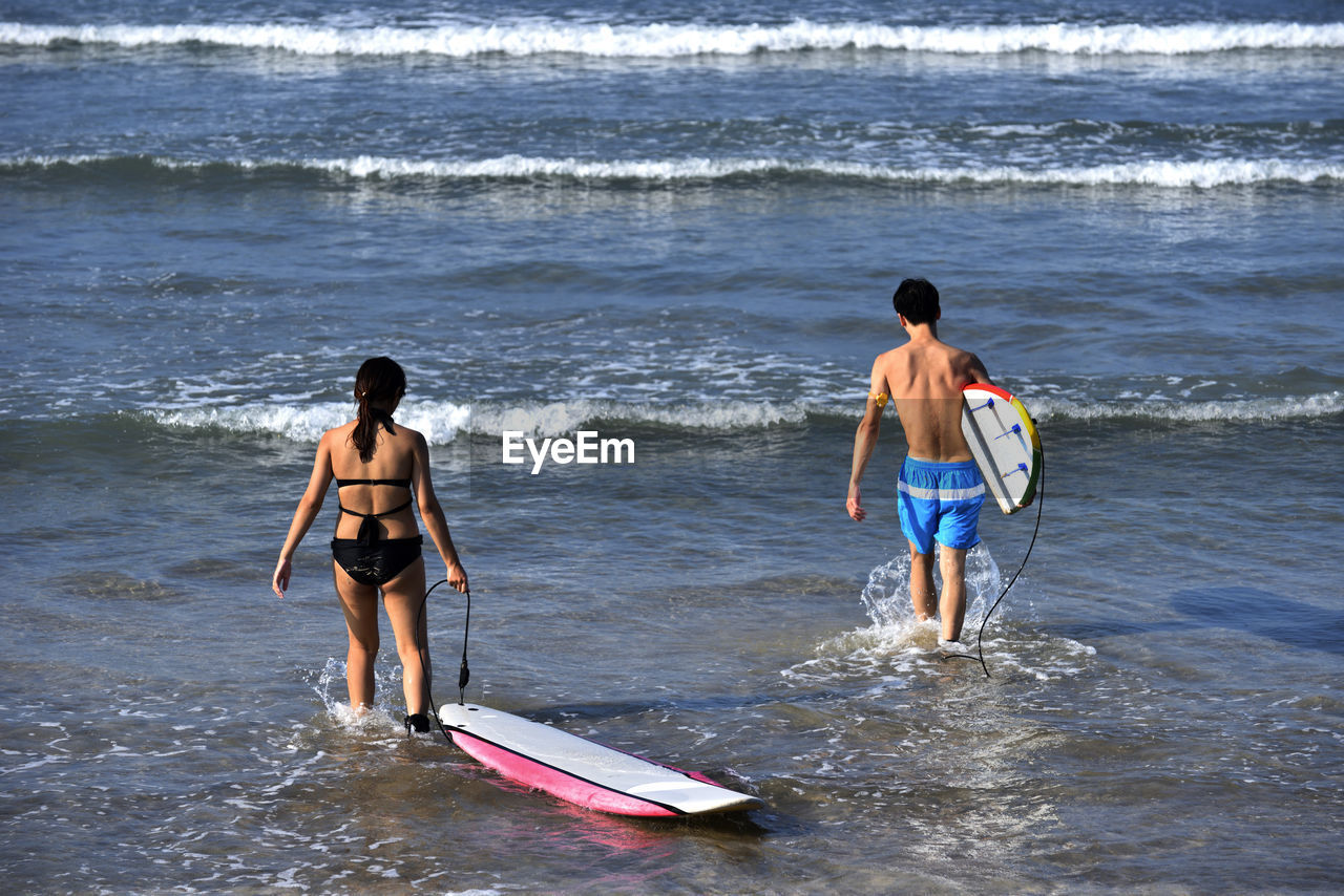 Rear view of friends holding surfboard while walking in sea