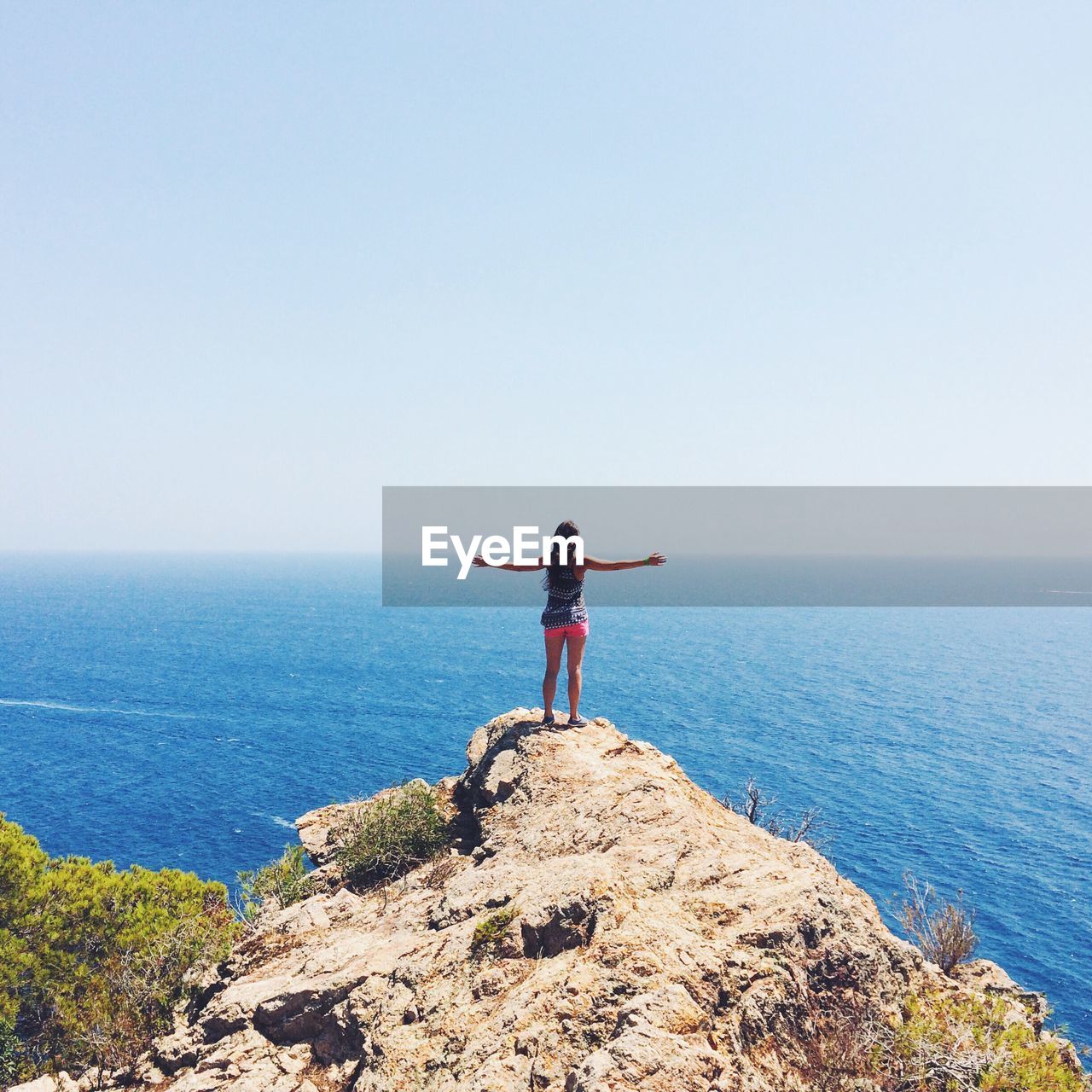 Rear view of woman standing on cliff while looking at sea against clear sky