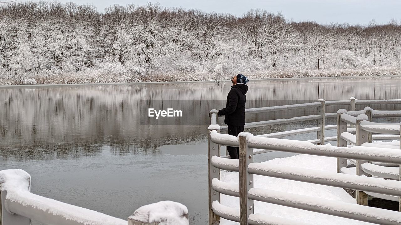 winter, snow, water, one person, nature, lake, cold temperature, railing, adult, tranquility, tranquil scene, beauty in nature, day, lifestyles, scenics - nature, full length, leisure activity, women, warm clothing, men, person, outdoors, clothing, tree, sky, wood, non-urban scene, environment, plant, reflection, landscape, ice, standing, female, relaxation