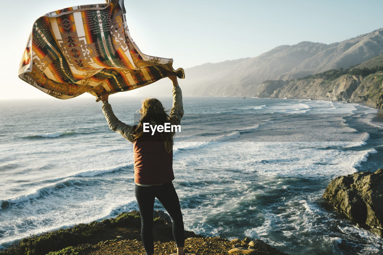 Rear view of woman holding scarf while standing on rock against sea