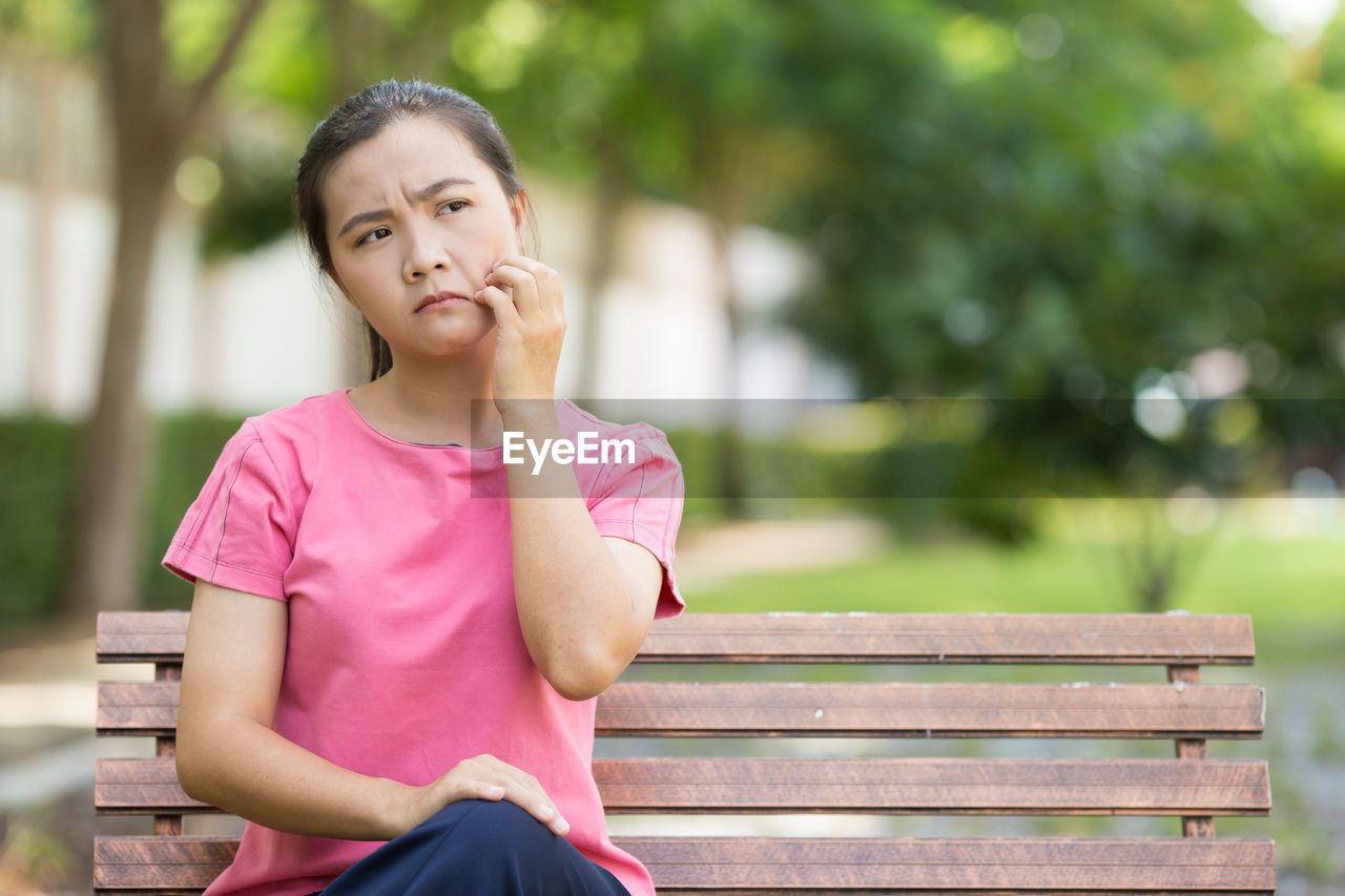 Young woman scratching cheek while sitting on bench at park