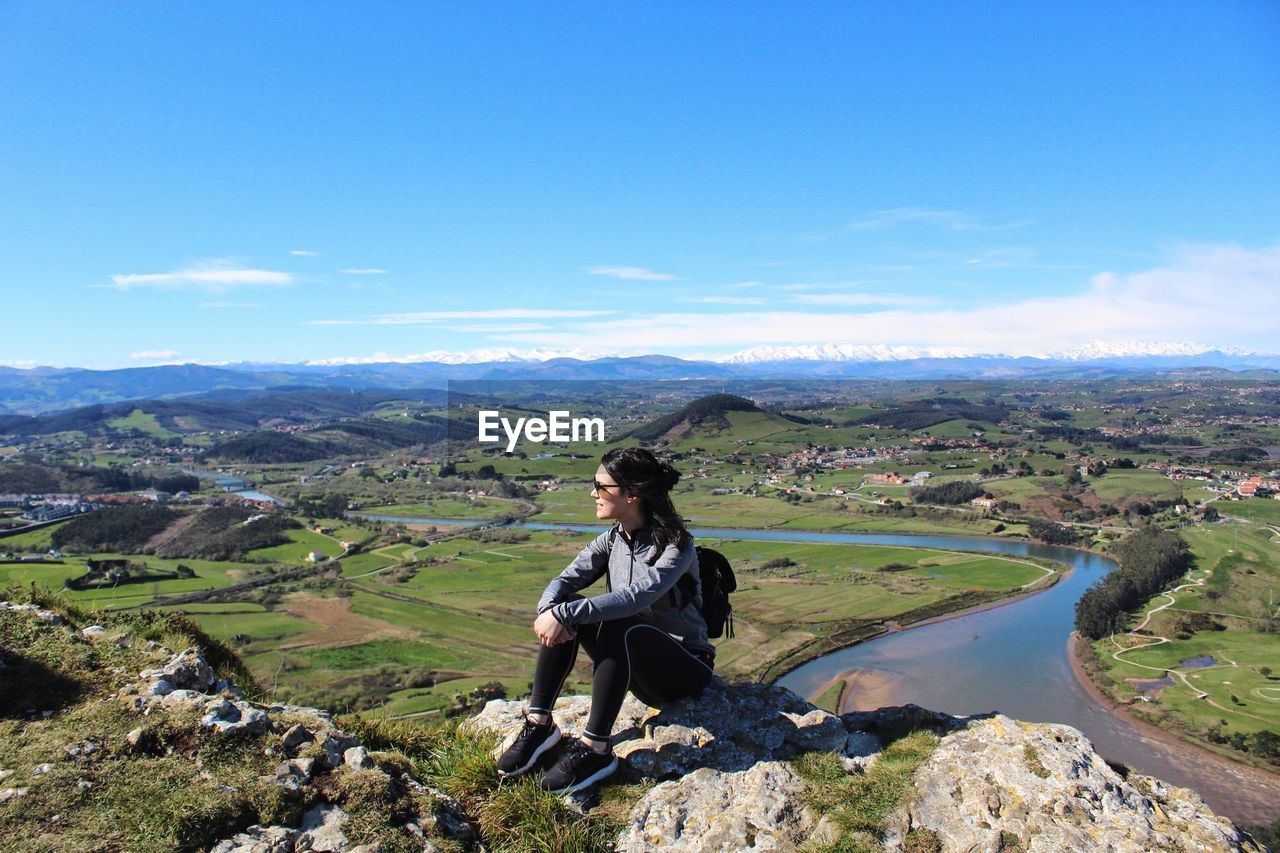 Young woman wearing sunglasses sitting on mountain against blue sky during sunny day