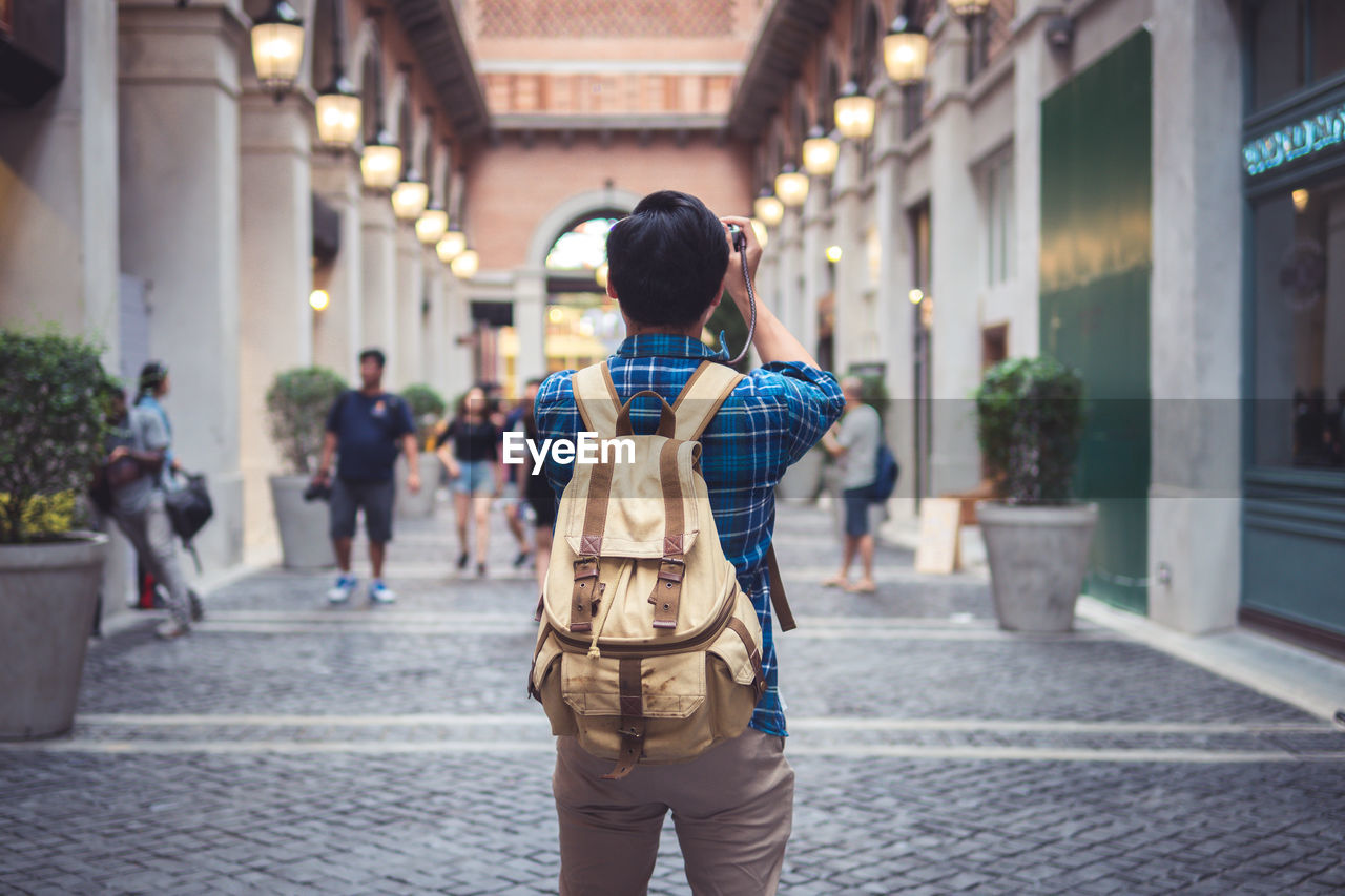 Rear view of tourist with backpack photographing on street in city