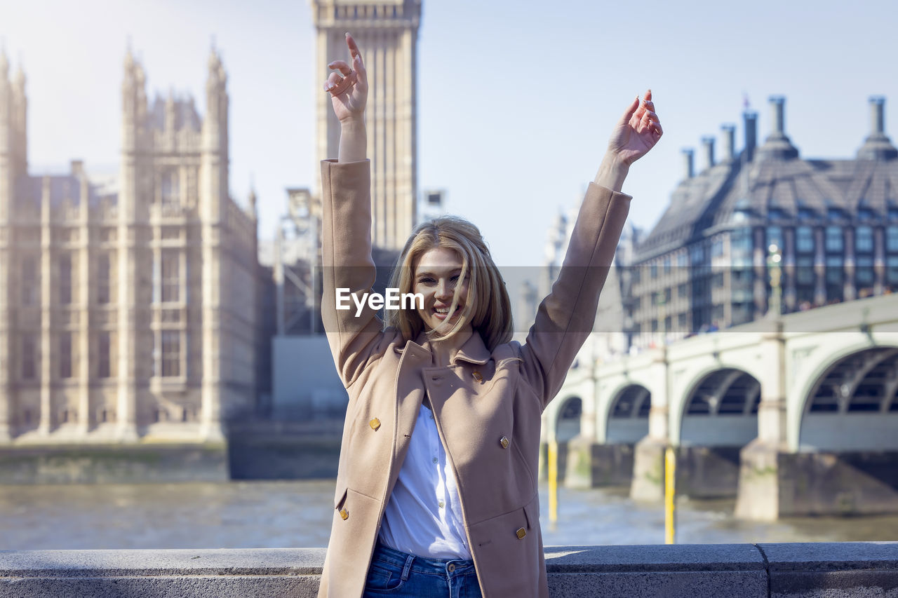 Portrait of young woman with arms raised standing against bridge