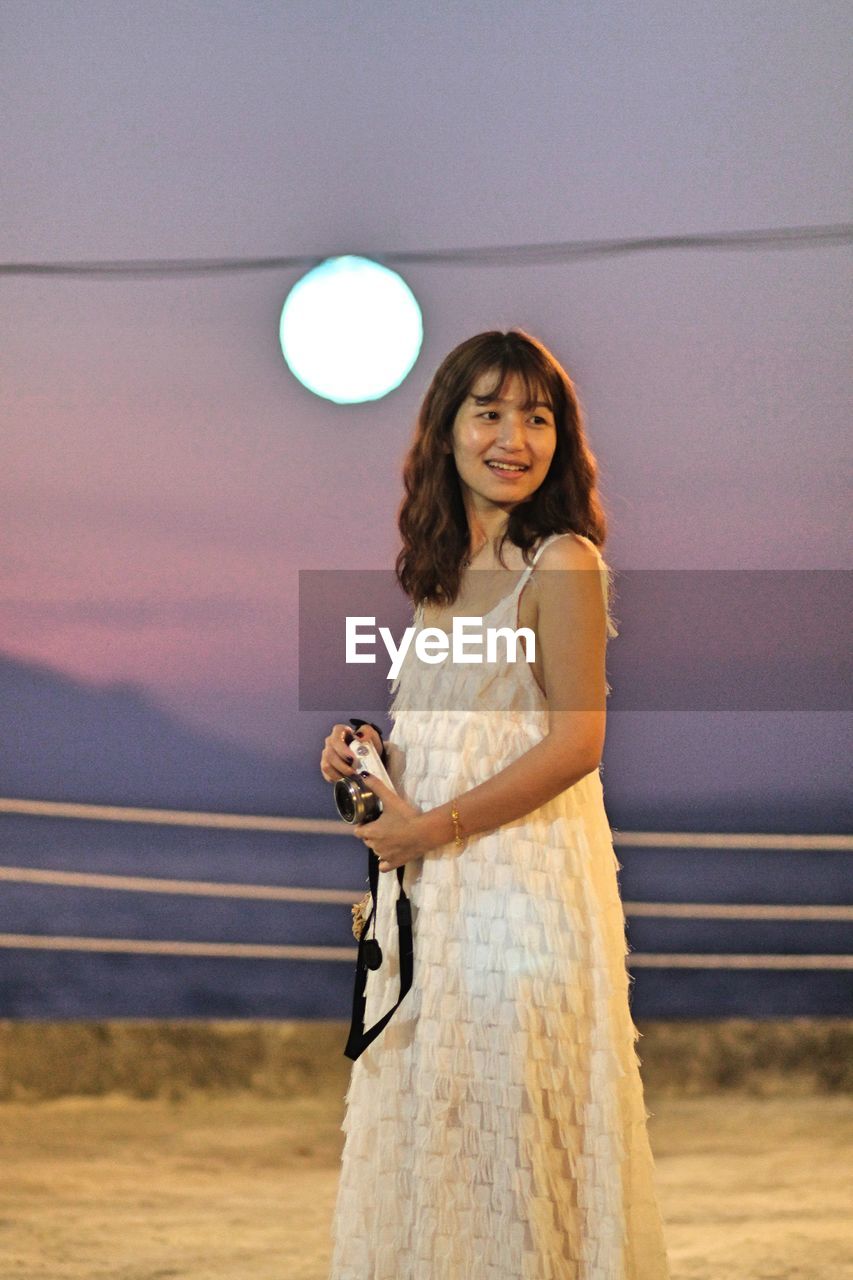 Smiling young woman holding camera while standing on land against sky at night