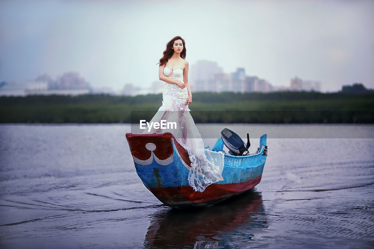 Beautiful female model in white evening gown standing on boat on shore