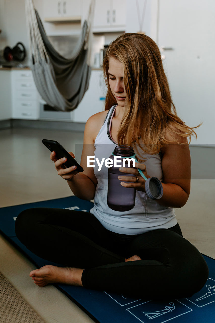 Smiling young female in sportswear holding water bottle and taking selfie on modern mobile phone during home workout