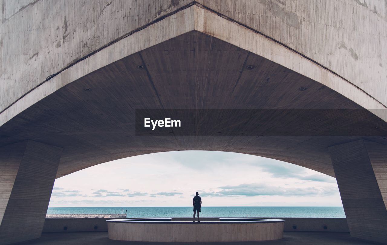 Rear view of man standing under bridge by sea