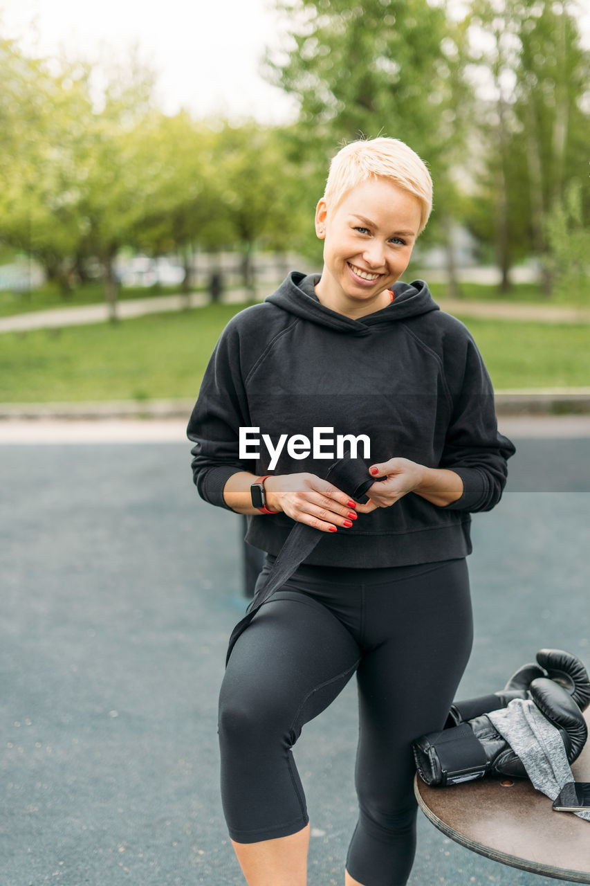 Young athletic blonde woman shakes boxing bandages on hands before training on the park