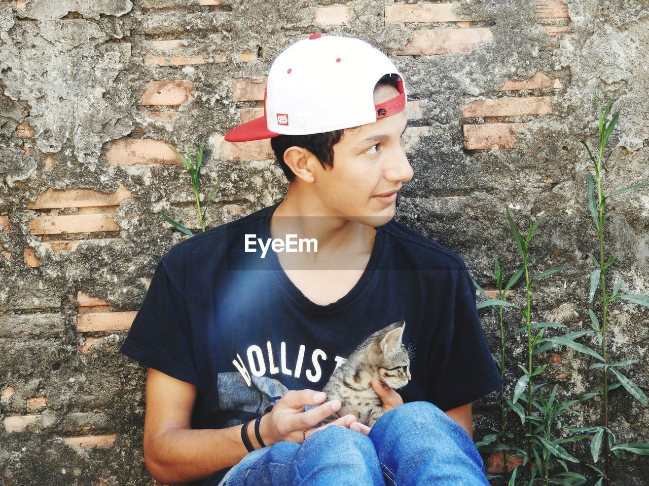 Teenage boy with cat looking away while sitting against wall