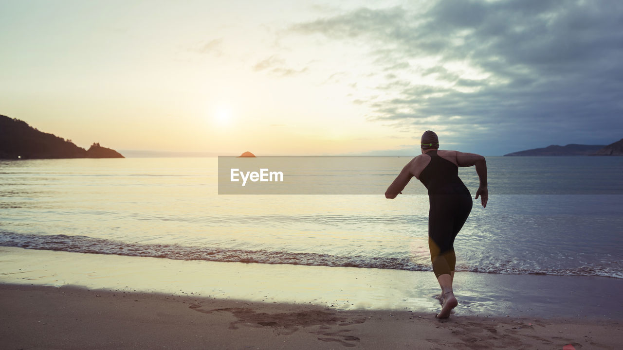 Back view of unrecognizable professional swimmer in swimsuit and cap ready for running in sea water during training at seaside at sunset