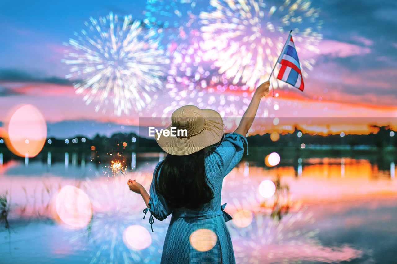 Rear view of woman with thai flag standing by lake against fireworks