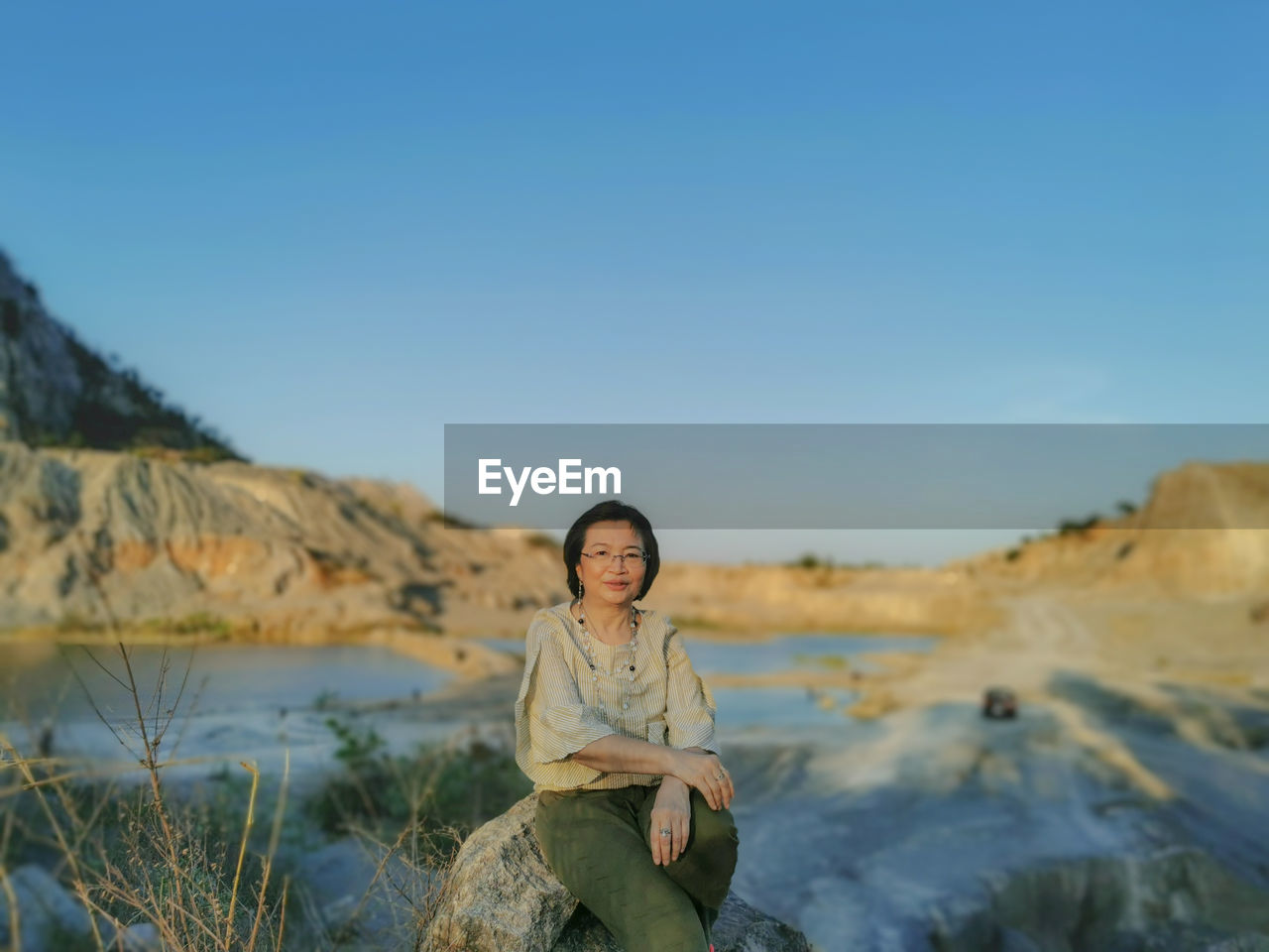 Portrait of smiling young woman sitting on rock against sky