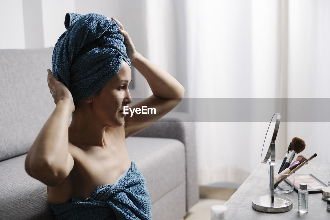Side view of young bare shouldered female with bath towel on head sitting near table with cosmetics and mirror and preparing for beauty procedure at home