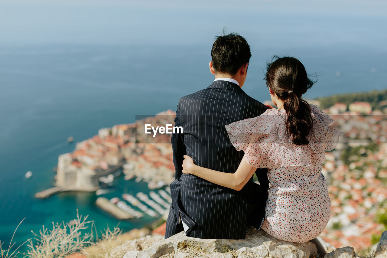 Rear view of married couple sitting on rock against sea