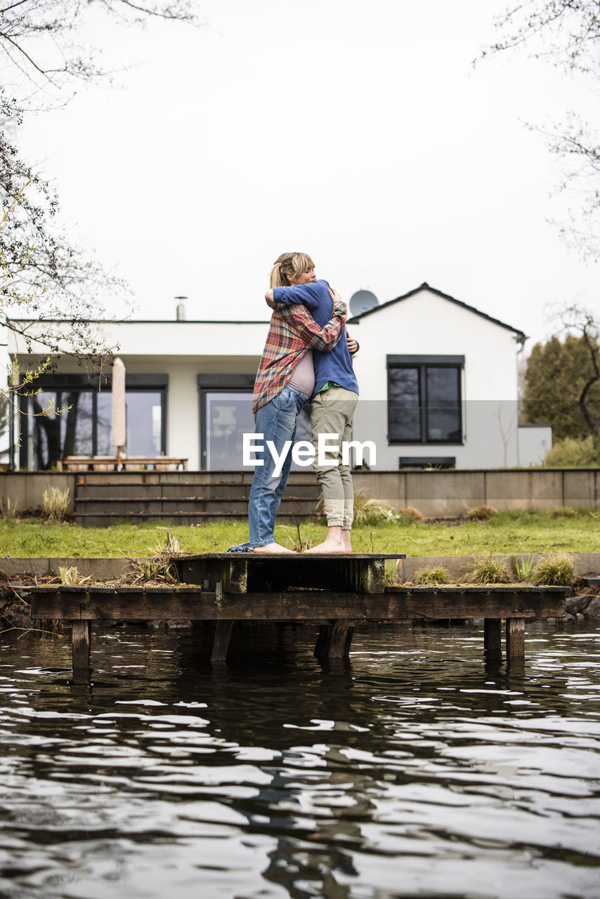 Expectant couple embracing each other standing on jetty in front of house