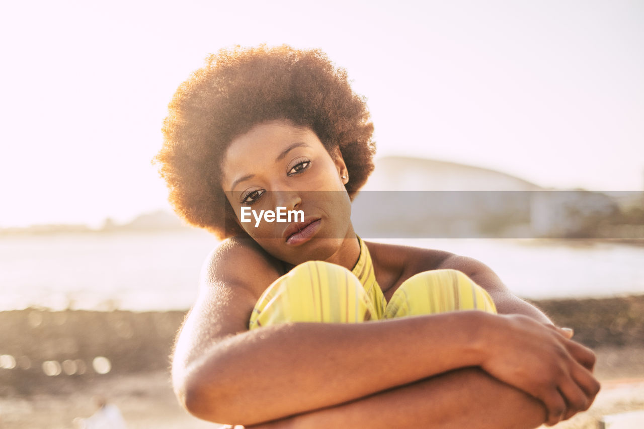 Portrait of young woman hugging knees while sitting against sky during sunset