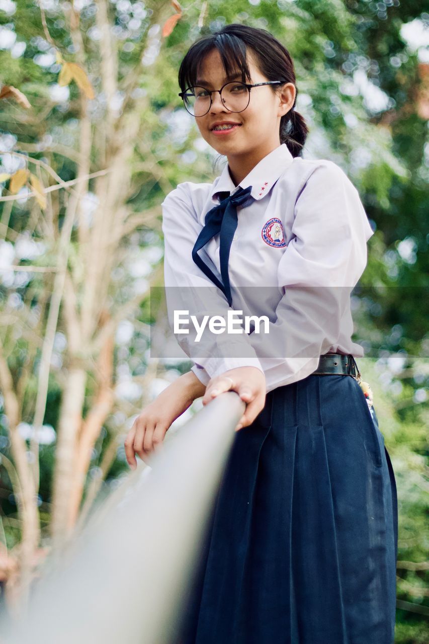 Low angle portrait of young woman wearing eyeglasses standing by railing against plants in park