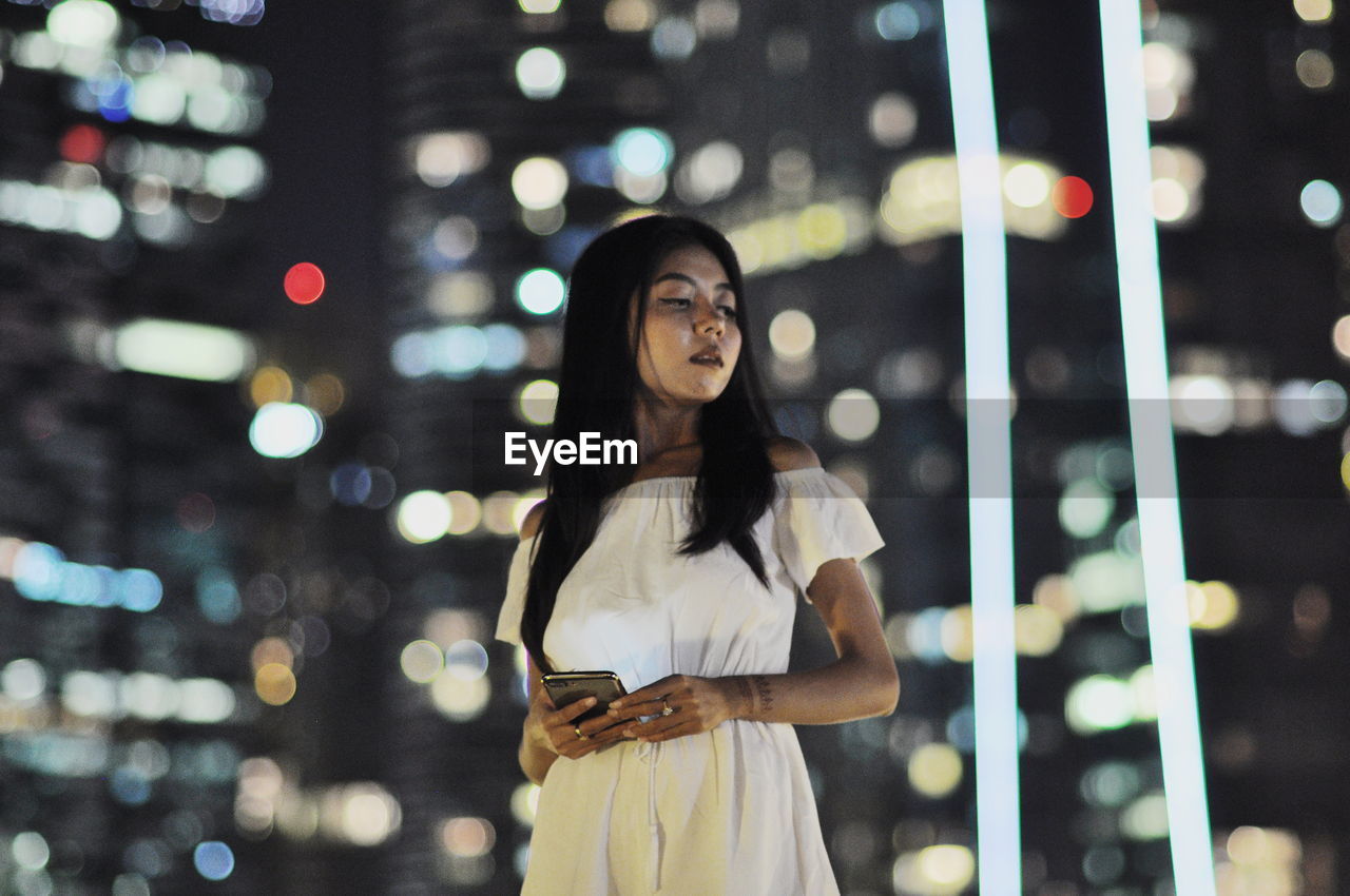 PORTRAIT OF SMILING YOUNG WOMAN STANDING AGAINST ILLUMINATED LIGHTS