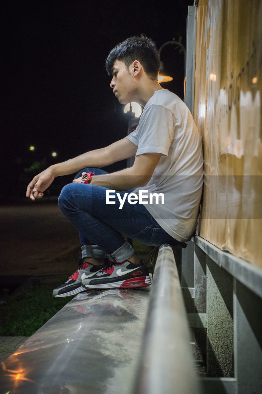 Side view of young man looking away while sitting against wall