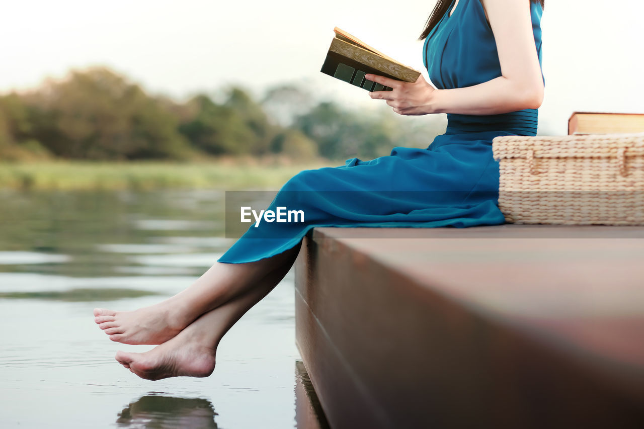Low section of mature woman reading book sitting on pier over lake