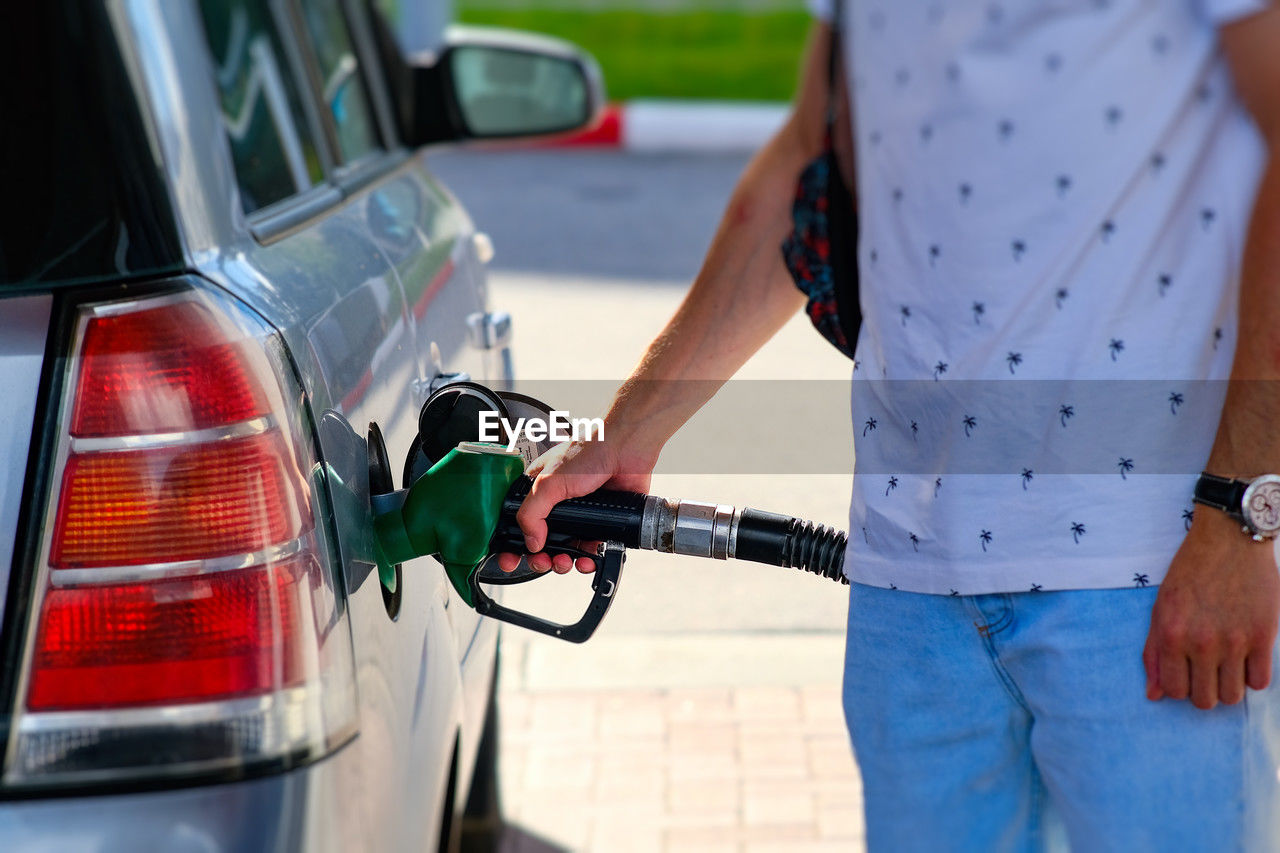 fuel pump, car, motor vehicle, mode of transportation, transportation, filling, gasoline, adult, gas station, power generation, one person, midsection, casual clothing, vehicle, fossil fuel, driving, day, standing, travel, land vehicle, holding, outdoors, focus on foreground
