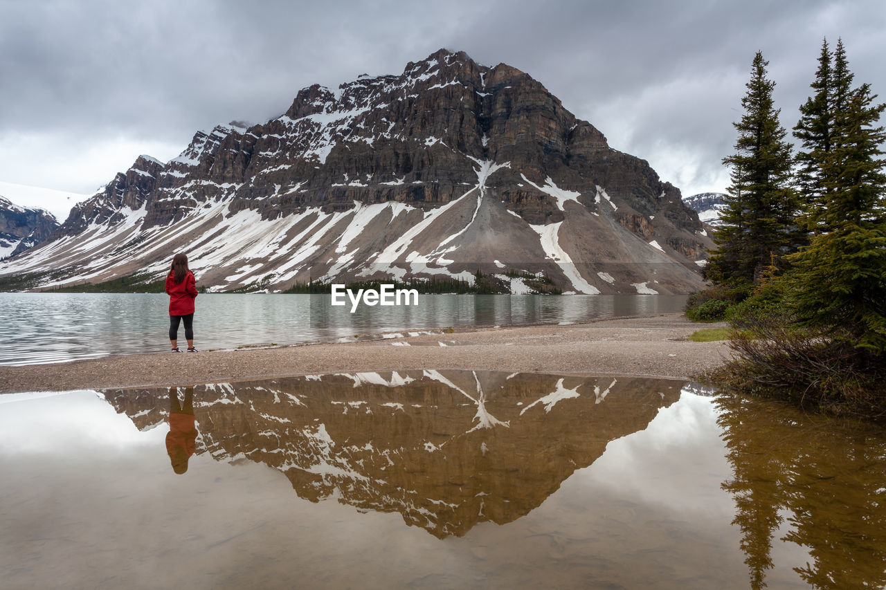 Rear view of woman standing at lakeshore against mountains during winter