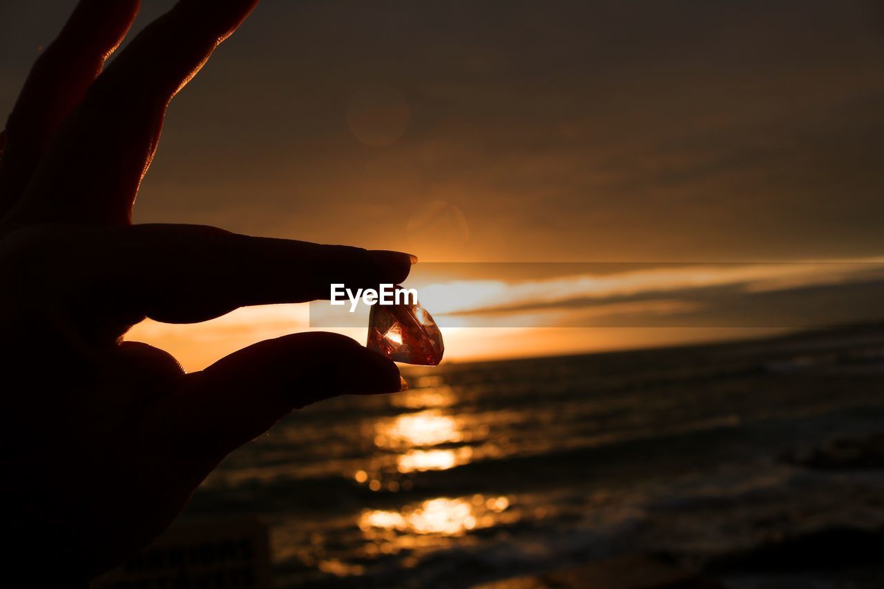Cropped hand of woman holding diamond against sea at sunset