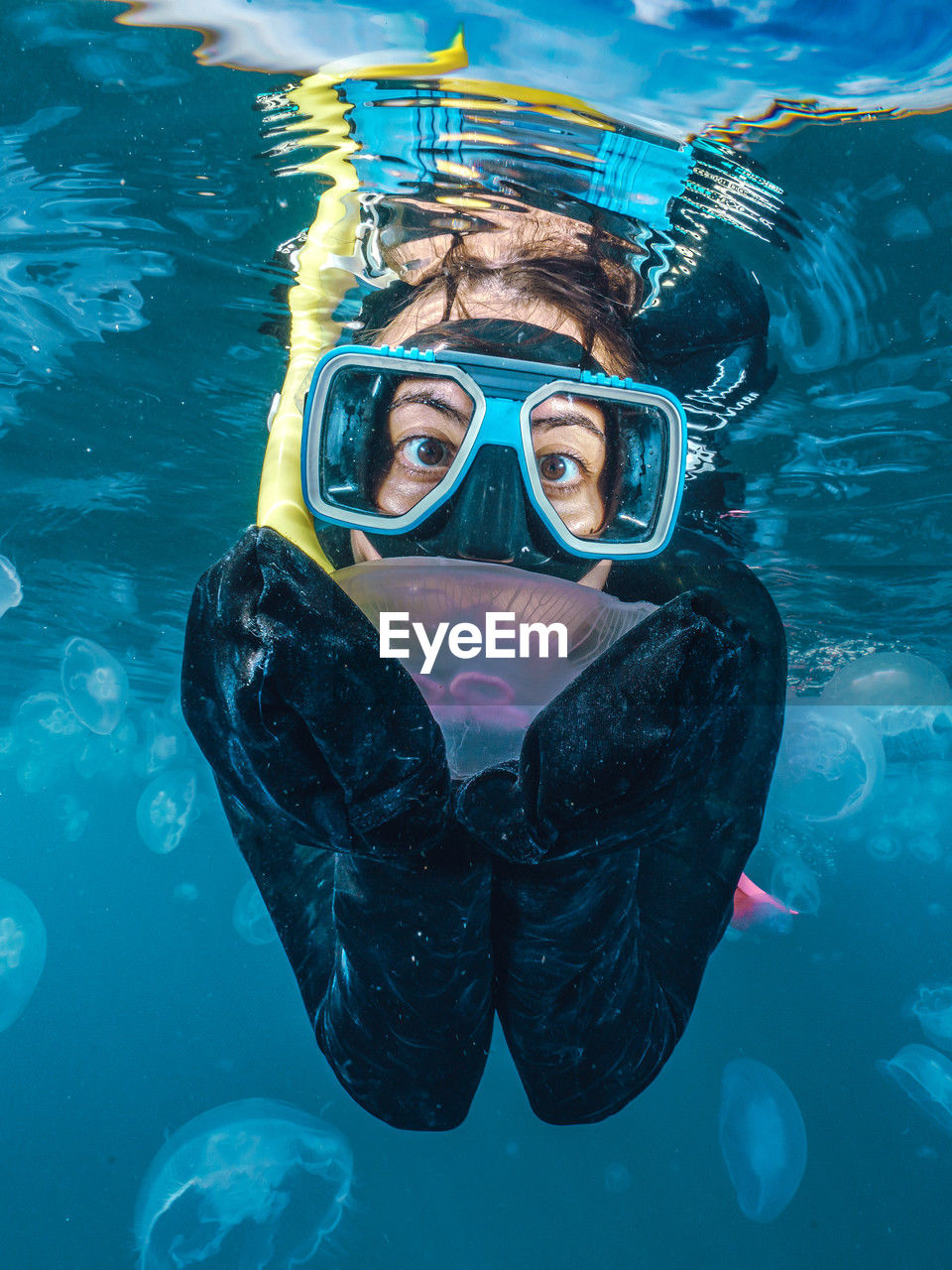 A woman in a black wetsuit is in the water with jellyfish. the jellyfish are floating around her