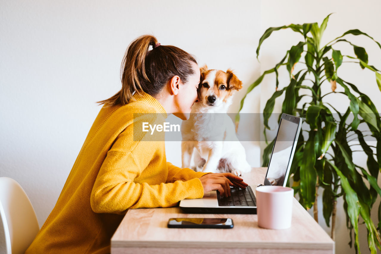 Side view of woman playing with dog while using laptop