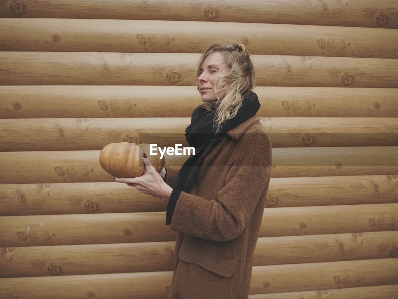 Side view of woman holding pumpkin while standing by wooden wall