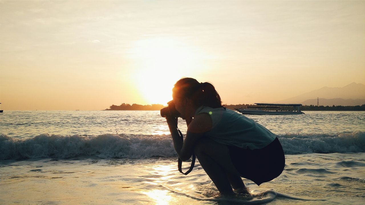 Woman crouching while photographing at beach during sunset
