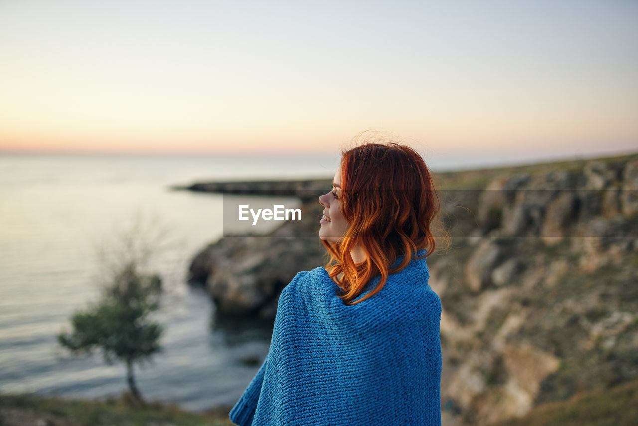 WOMAN LOOKING AT SEA SHORE AGAINST SUNSET SKY