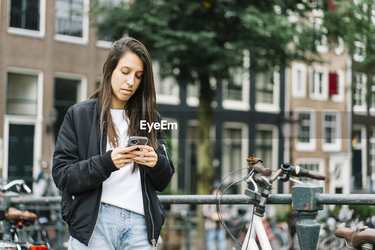 Side view of concentrated ethnic female traveler with long dark hair in casual clothes messaging on mobile phone while standing near parked bicycle on bridge during trip in amsterdam