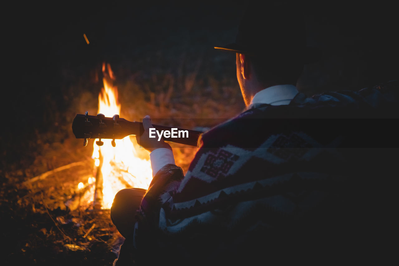 Rear view of man with guitar sitting by bonfire in forest