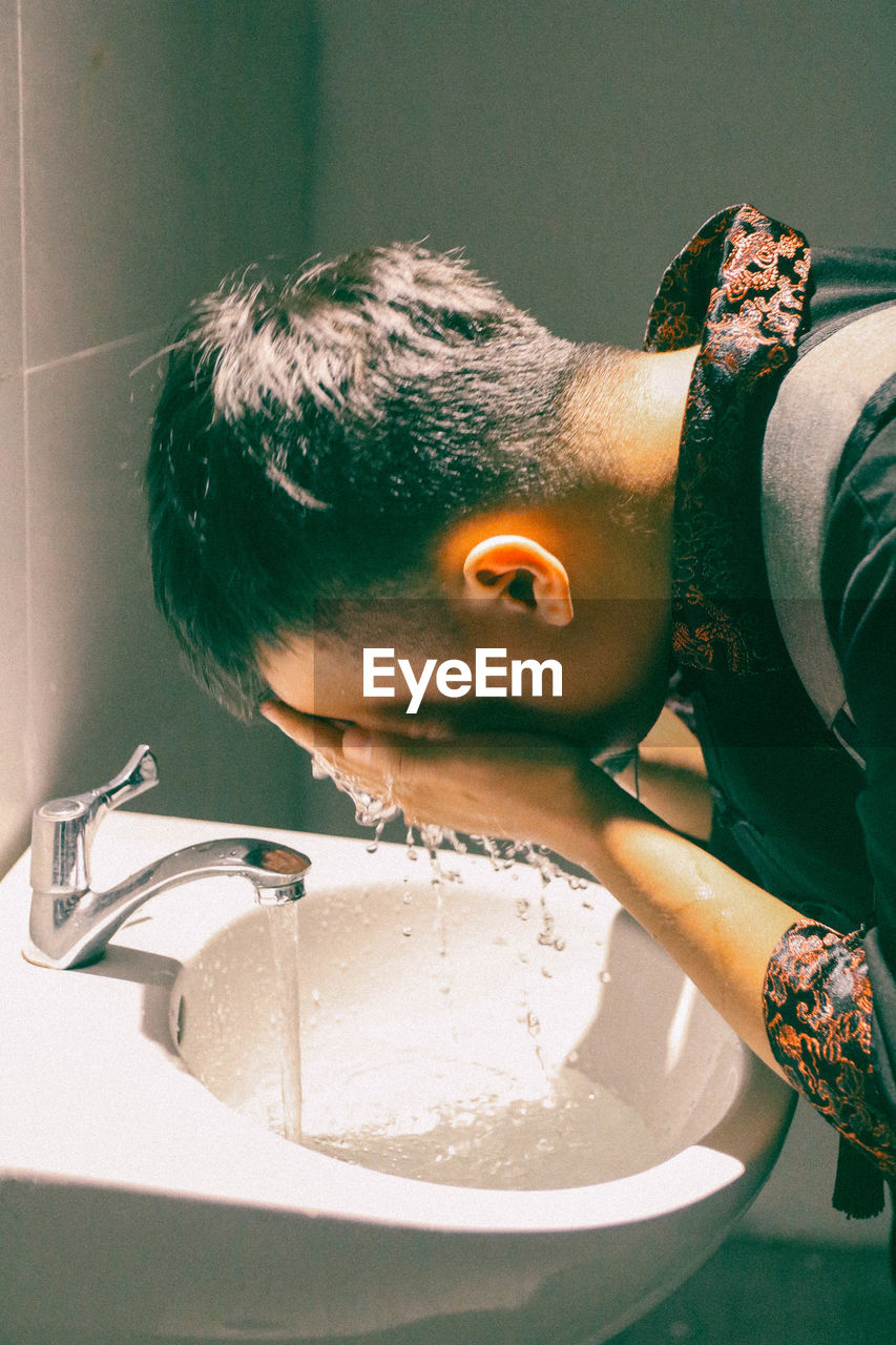 Side view of man washing face in bathroom sink