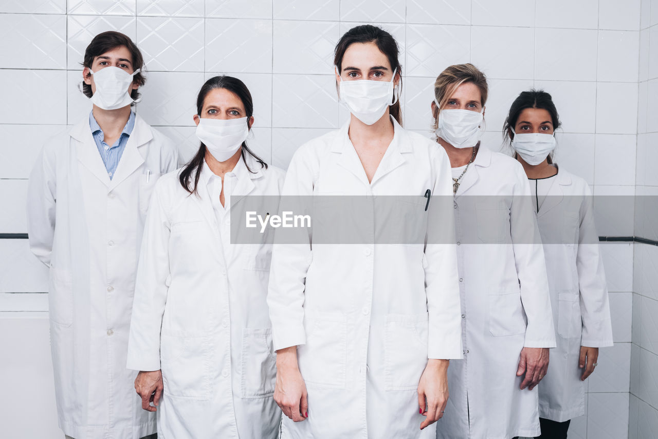 A group of five doctors with a mask on their face and a white coat standing up in a hospital room