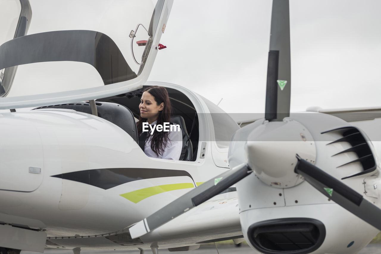 Smiling female pilot sitting in airplane against cloudy sky at airport