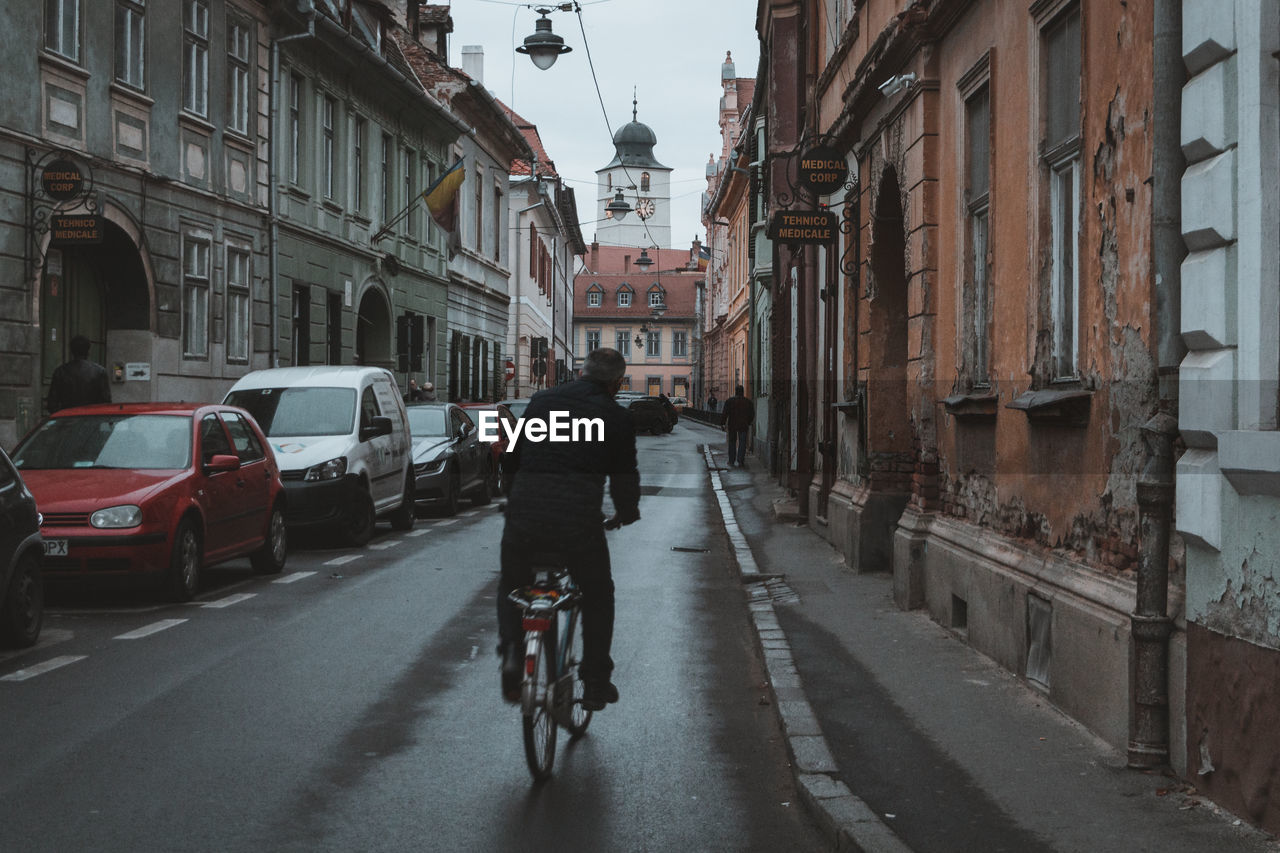 REAR VIEW OF MAN RIDING BICYCLES ON STREET AMIDST BUILDINGS