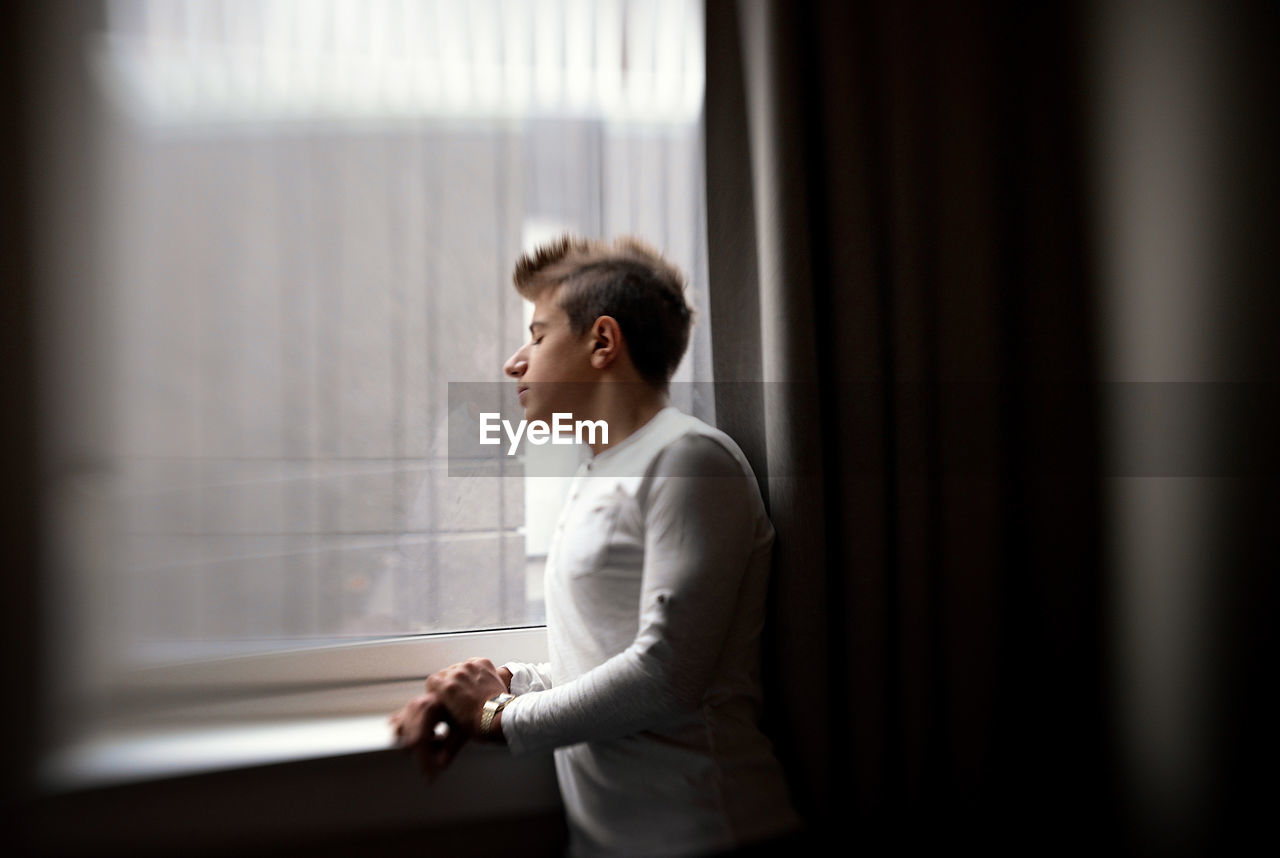 Young man standing by glass window at home