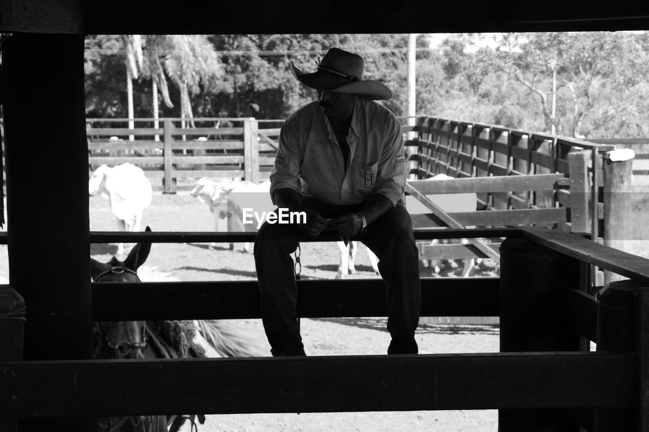 Full length of man wearing cowboy hat while sitting on fence at farm