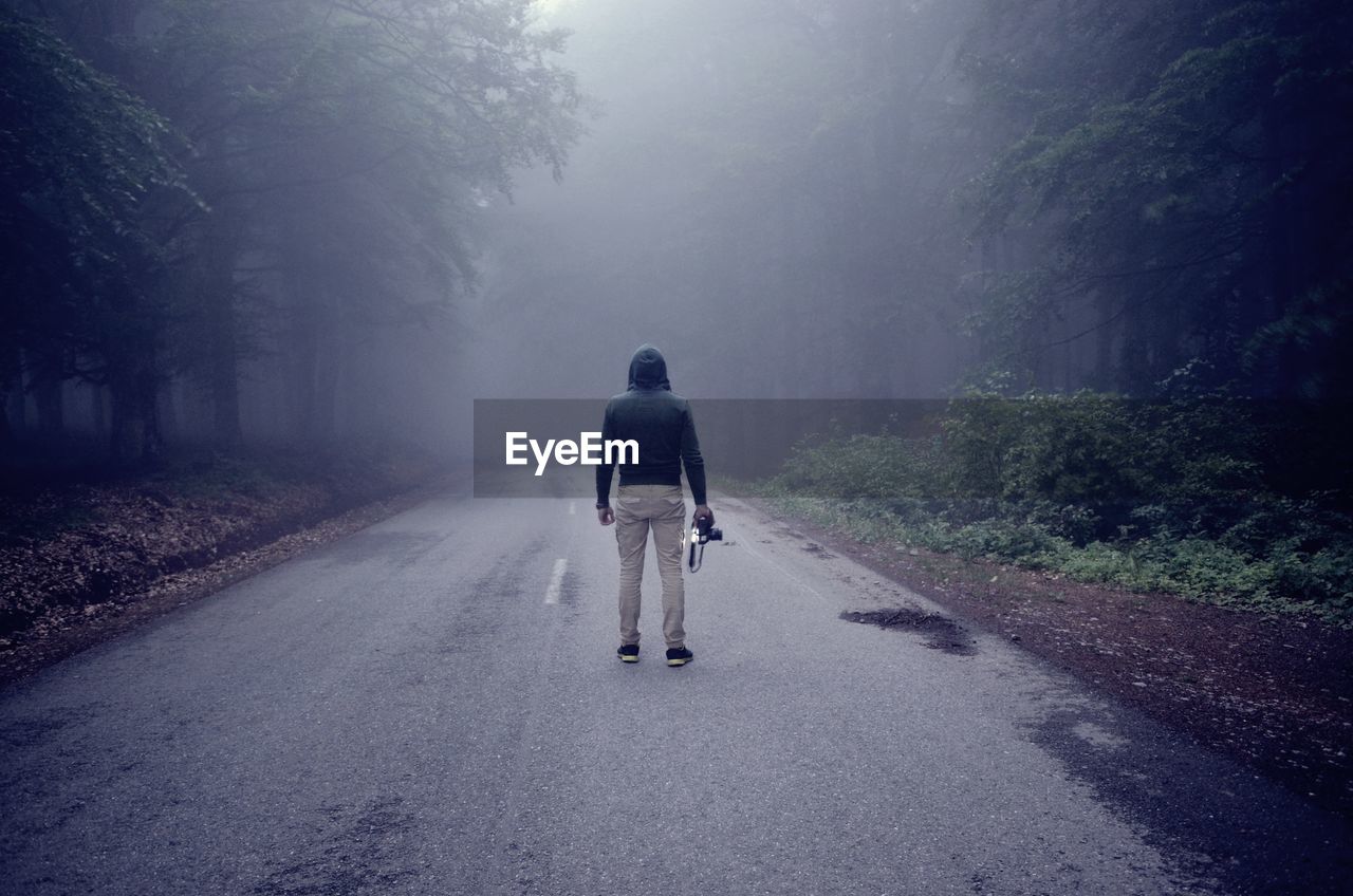 Rear view of man holding camera while standing on road amidst trees during foggy weather