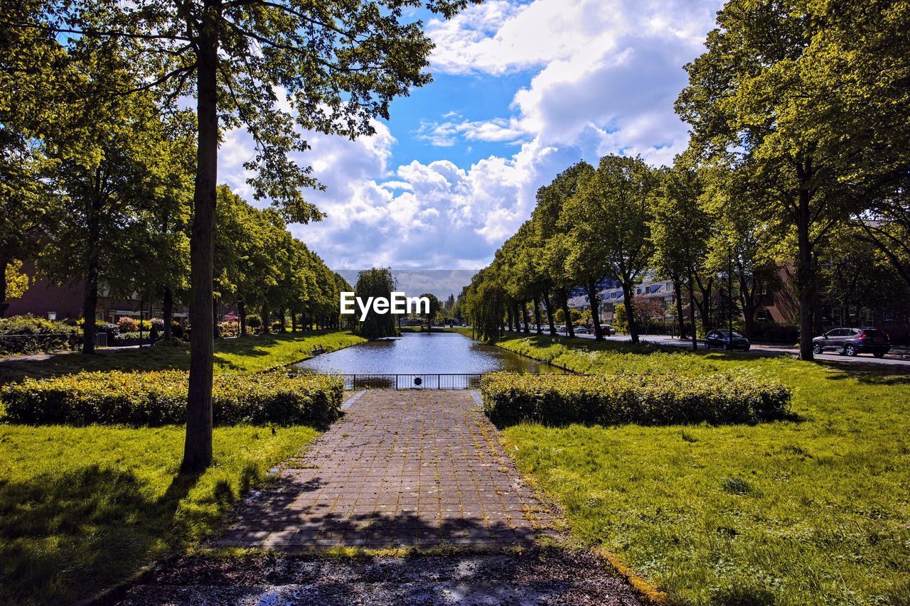 Footpath amidst trees in park with pond against sky