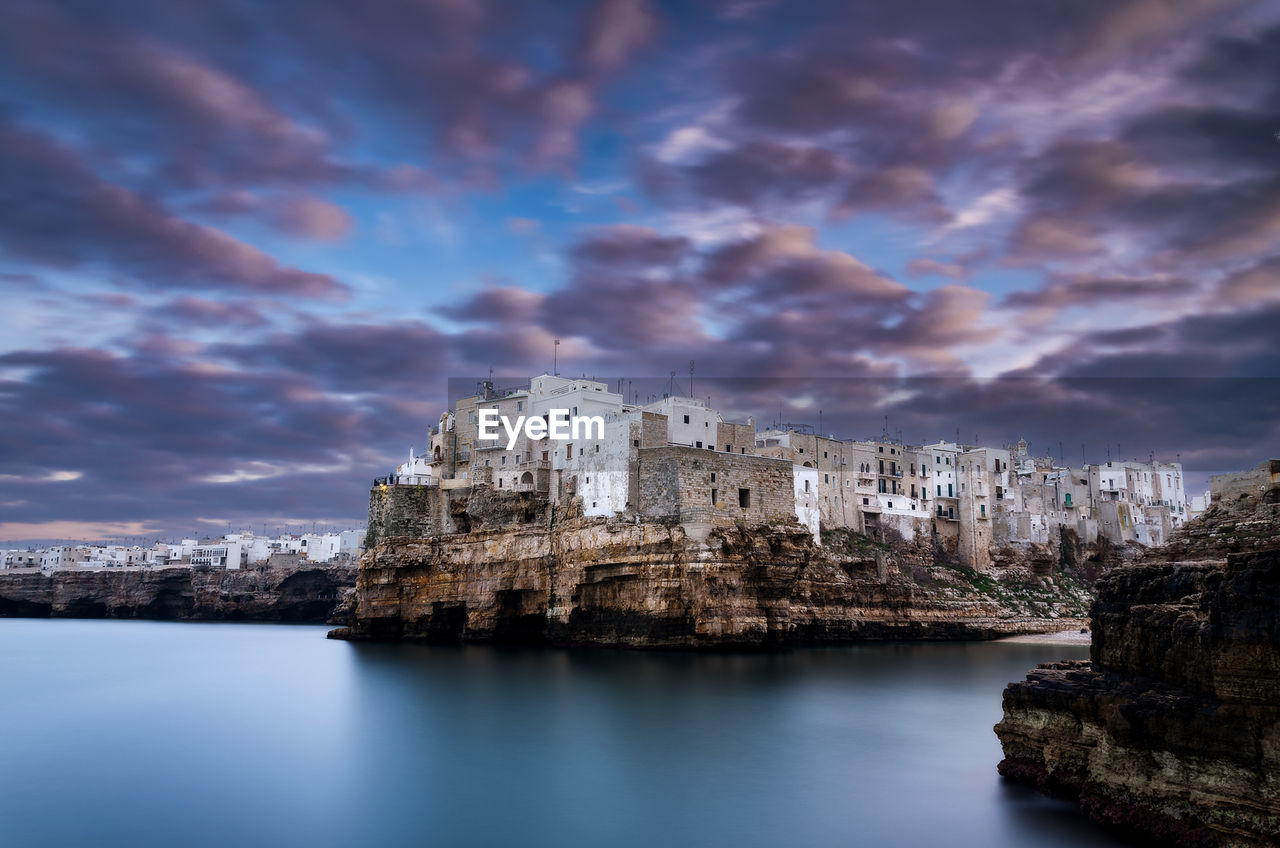 Buildings by sea against cloudy sky at dusk