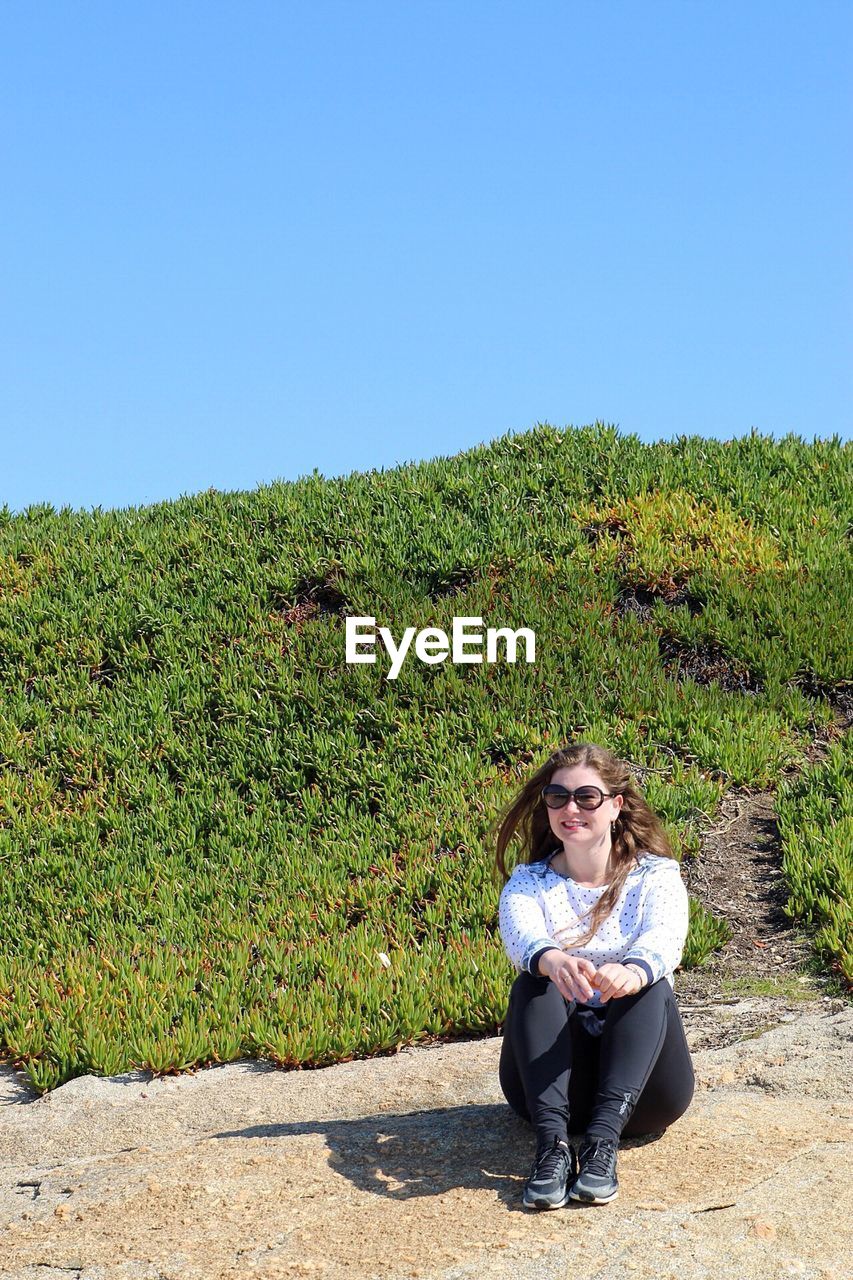 Portrait of woman sitting on footpath against clear blue sky