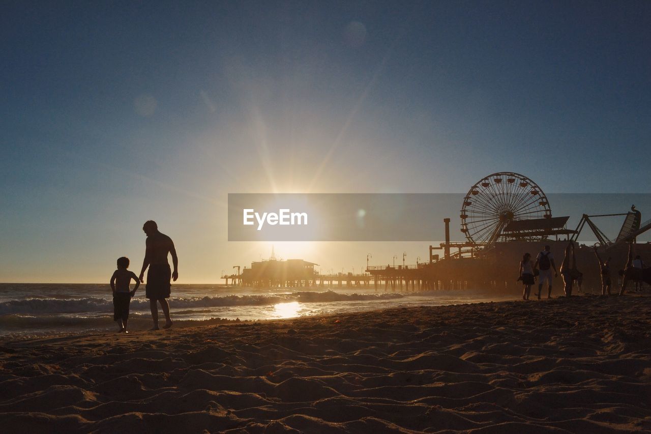 People walking at beach by santa monica pier against clear sky during sunset